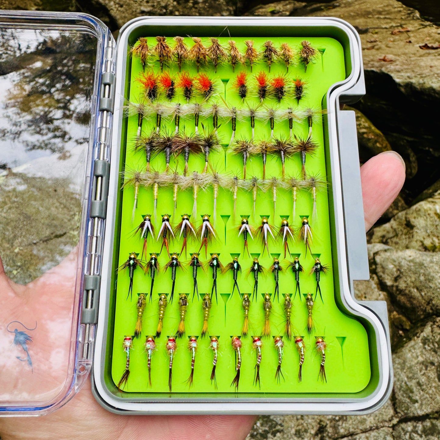 A lifestyle photo of The Essentials Fly Box from Fly Life Outdoors, open in a hand with a trout stream in the background.