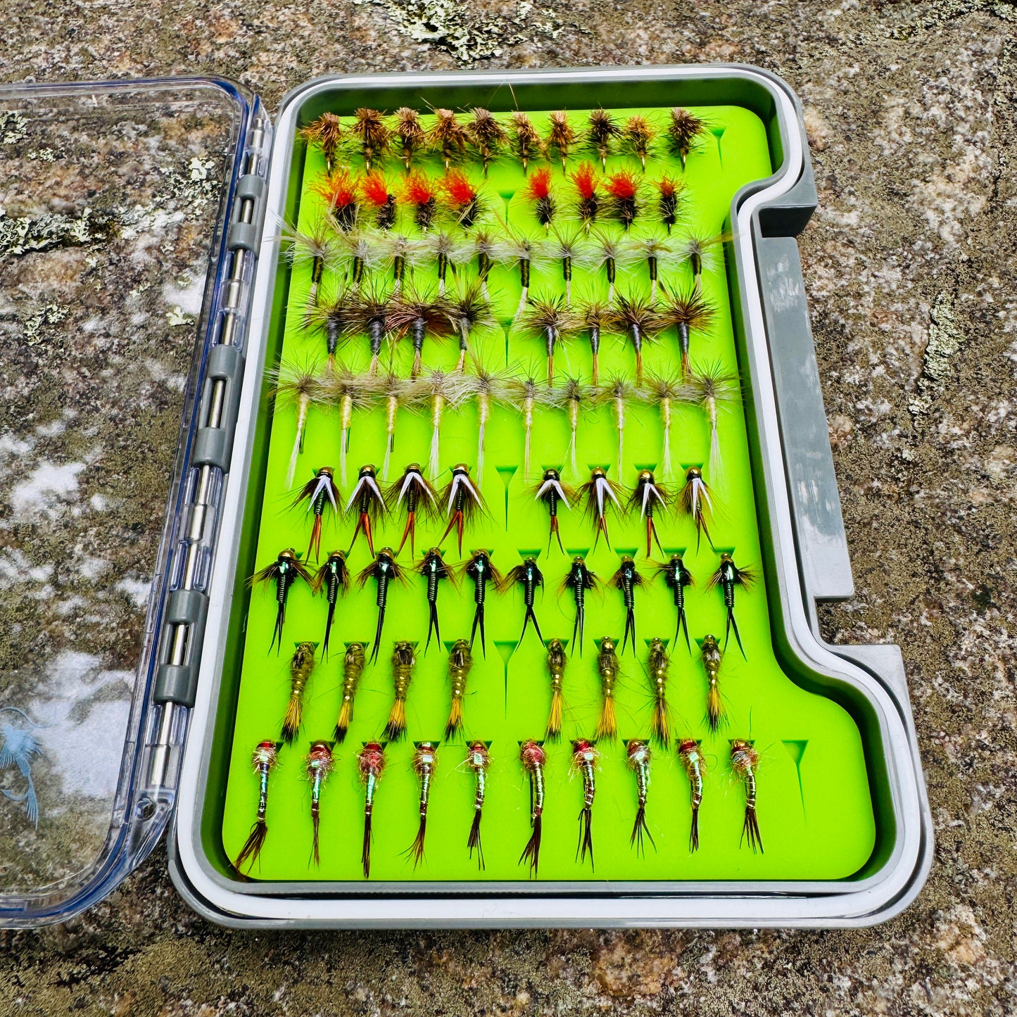 The complete Essentials Fly Box kit, open on a rock to show the full assortment of 82 hand-tied dry flies and nymphs and the green silicone insert that keeps them secure.
