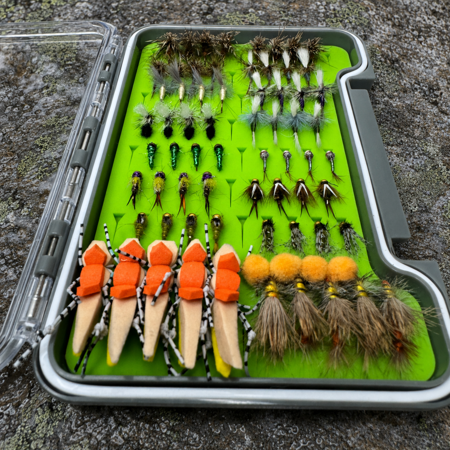 All-Water Arsenal loaded fly box angled view on granite river rock displaying 70 hand-tied dry flies, nymphs, and stonefly patterns for North Carolina trout fishing