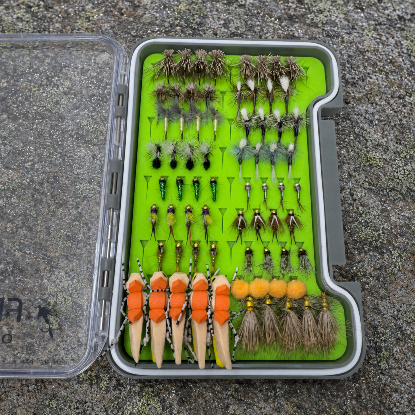 All-Water Arsenal loaded fly box open on river rock showing hand-tied NC trout flies including Elk Hair Caddis, Parachute Adams, Zebra Midges, Copper Johns, and foam stonefly patterns