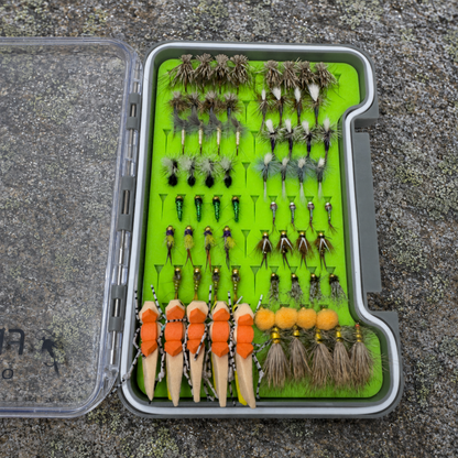 All-Water Arsenal loaded fly box open on river rock showing hand-tied NC trout flies including Elk Hair Caddis, Parachute Adams, Zebra Midges, Copper Johns, and foam stonefly patterns