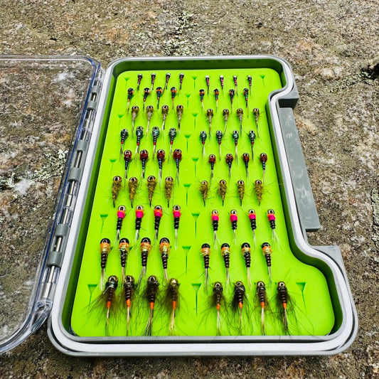 Depth Charge Fly Box open on granite rock, loaded with rows of beadhead nymphs in gold, copper, green, and pink bead patterns on green foam insert
