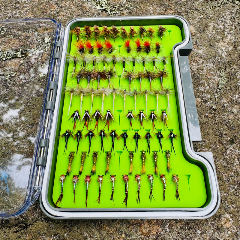 Essentials Fly Box open on granite rock showing rows of hand-tied dry flies, wet flies, and nymphs including parachute patterns and elk hair caddis on green foam insert