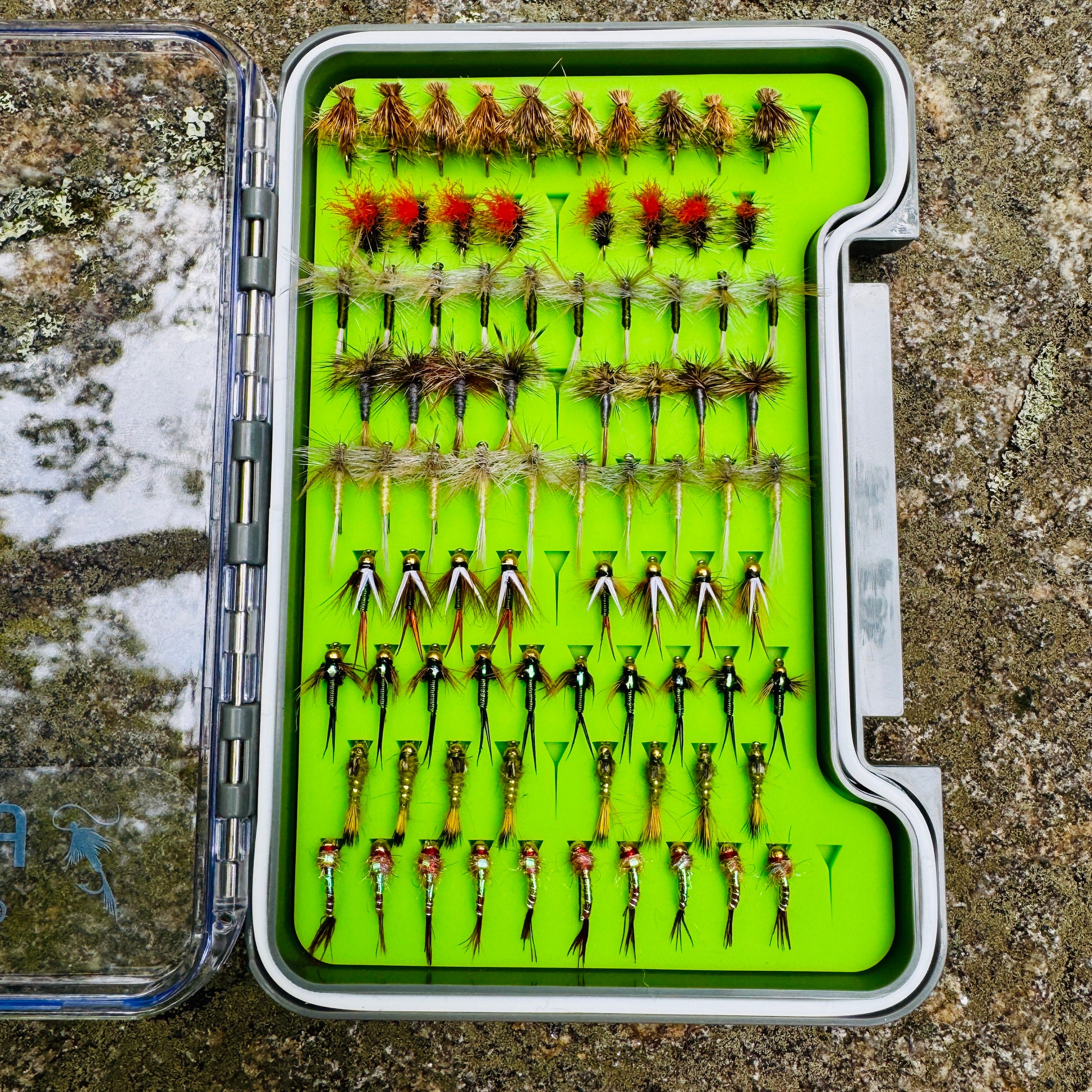 The complete Essentials Fly Box kit, open on a rock to show the full assortment of 82 hand-tied dry flies and nymphs.