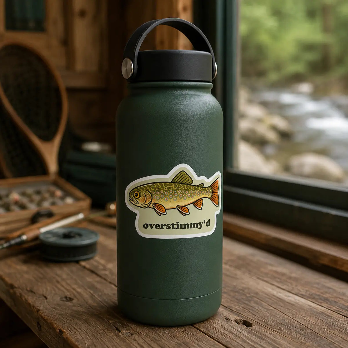 Overstimmy’d brook trout sticker on a green water bottle at a fly-tying bench overlooking a stream