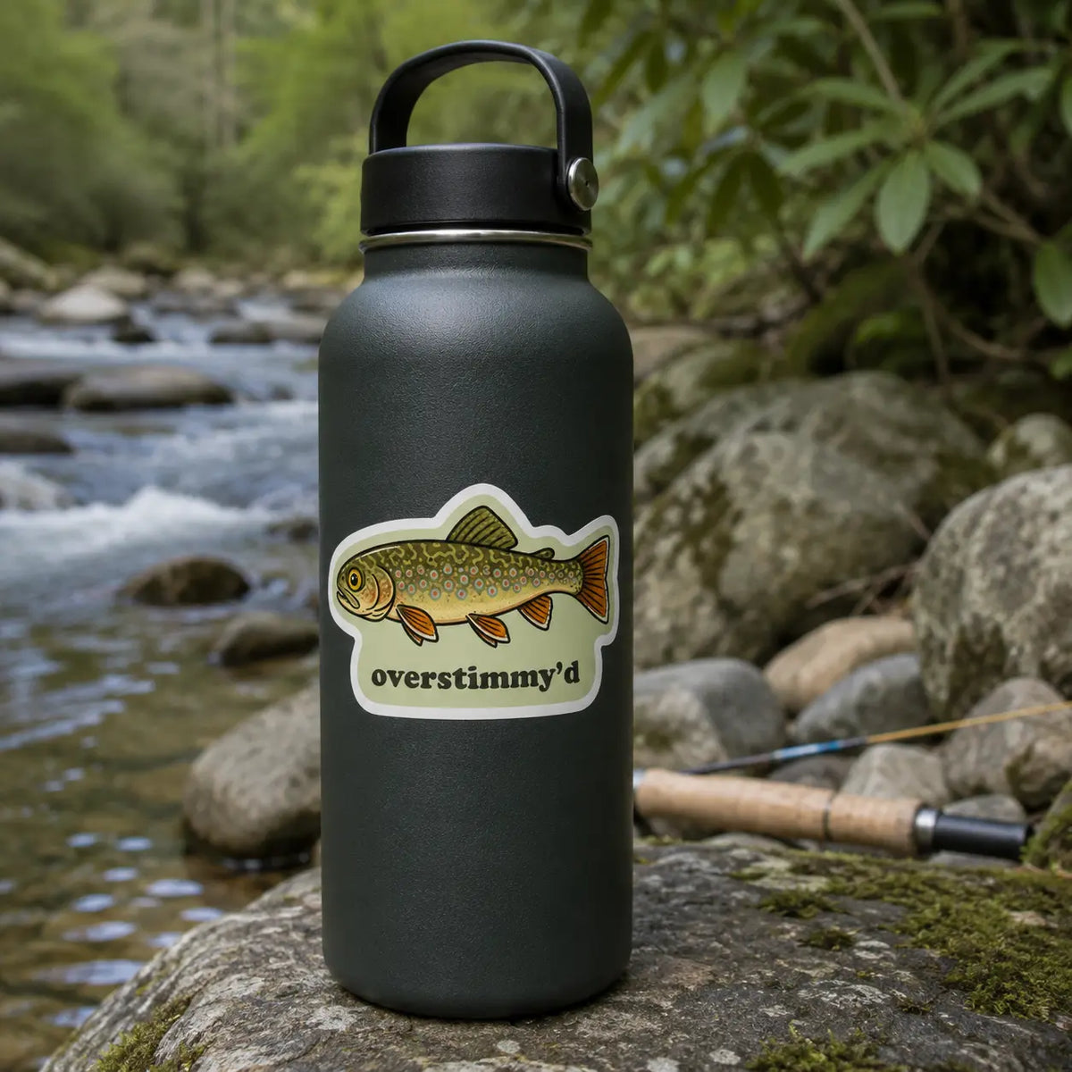 Overstimmy’d brook trout sticker on a black water bottle beside a mountain trout stream